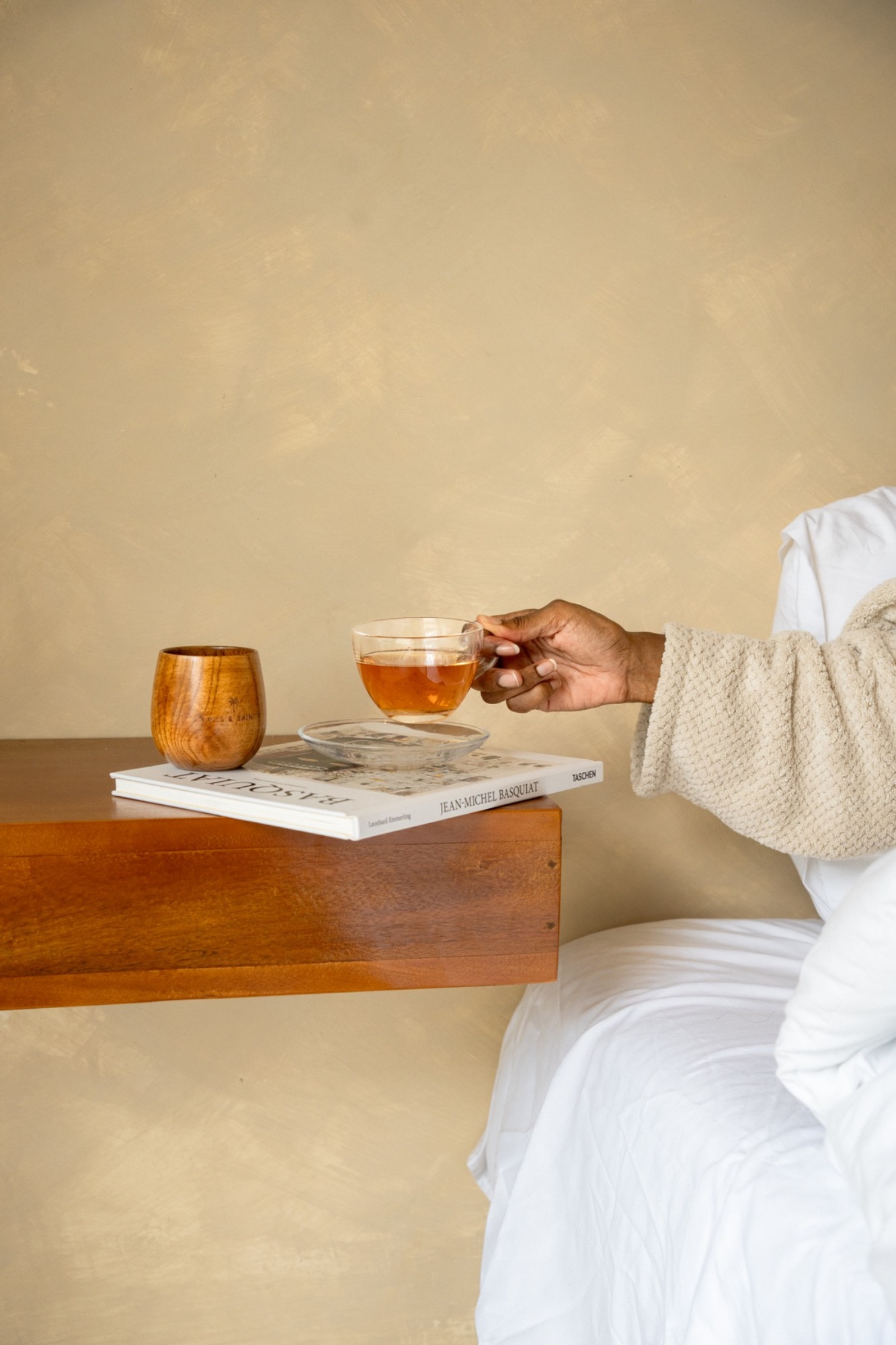 Wooden Yves and Saints Ahomka cup beside a book and a guest holding tea